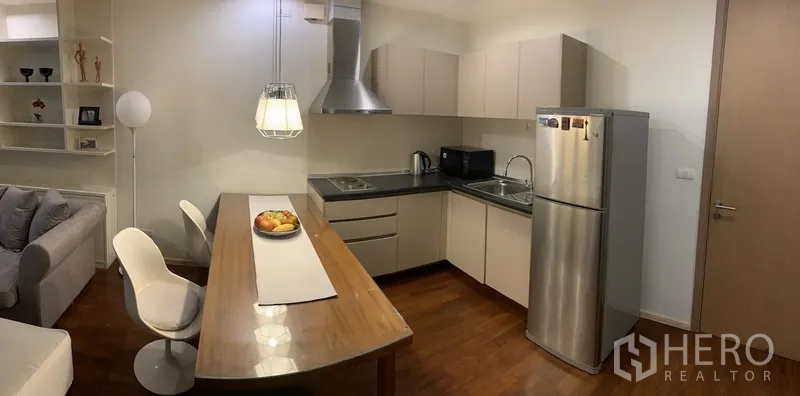 Condominium for rent or sale in Khlong Toei, Bangkok - Modern L-shaped kitchen with upper and lower cabinets, stainless refrigerator and hood, plus a wooden dining table for two.
