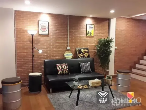 Townhouse for rent in Khlong Toei, Bangkok - Cozy living room featuring an exposed brick wall, black sofa, glass coffee table, and stairway lighting.