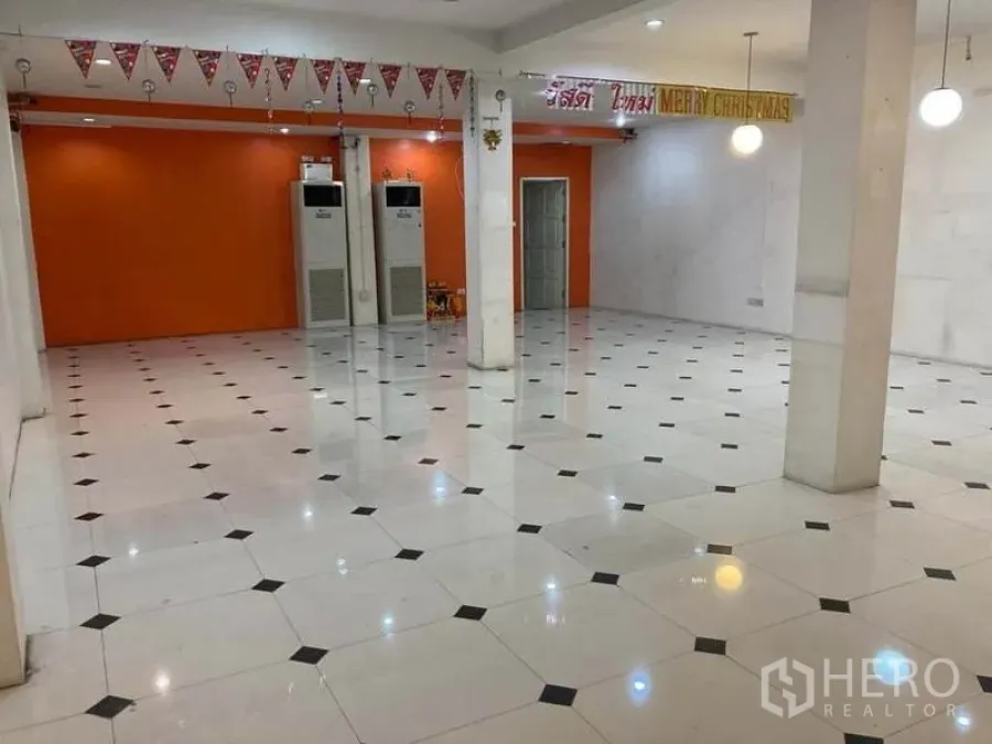 Shophouse for rent in Suan Luang, Bangkok - Large retail hall with an orange feature wall, pillars, ceiling lights and freestanding air-conditioning units.
