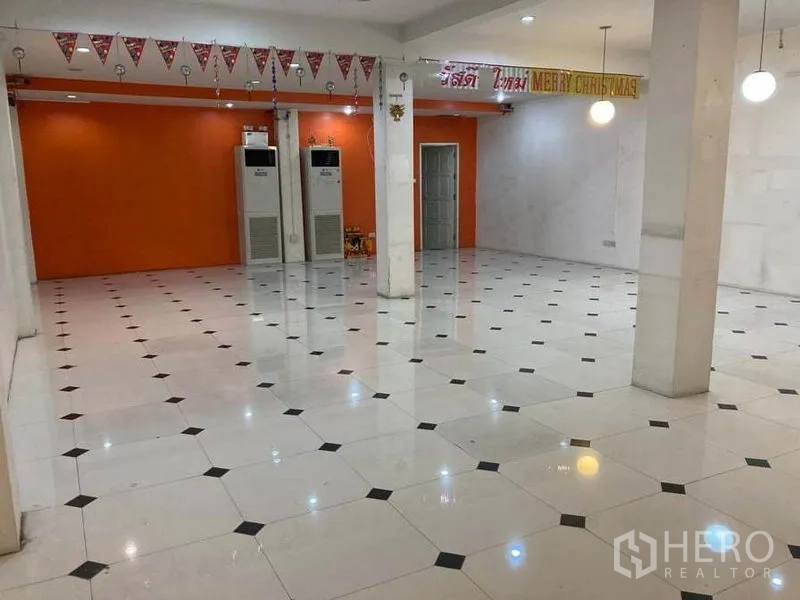 Shophouse for rent in Suan Luang, Bangkok - Large retail hall with an orange feature wall, pillars, ceiling lights and freestanding air-conditioning units.