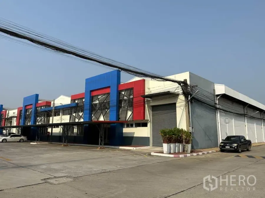 Factory for rent in Bang Phli, Samut Prakan - Angled view of modern factory exterior with blue and red facade, roller shutter door and parking bay.