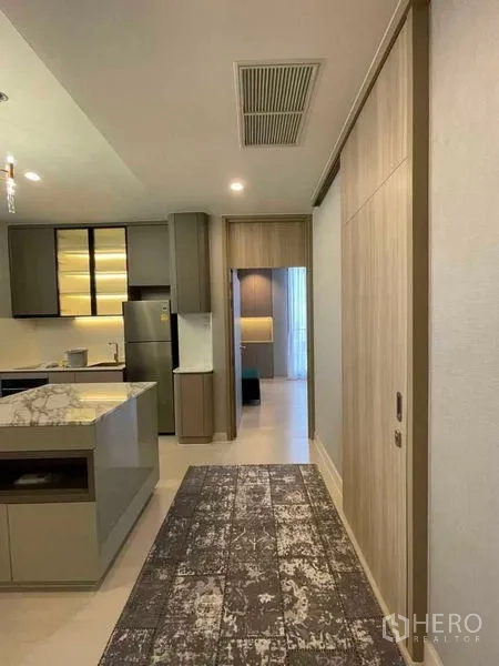 Condominium for rent in Pathum Wan, Bangkok - Hallway leading to an open kitchen with marble island, built-in cabinets and a stainless-steel fridge.