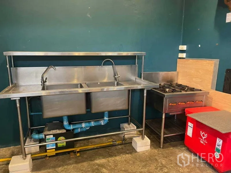 Retail Space for rent in Khlong Toei, Bangkok - Stainless-steel double sink with faucet beside a gas burner stove, visible plumbing and power outlets against a teal wall.