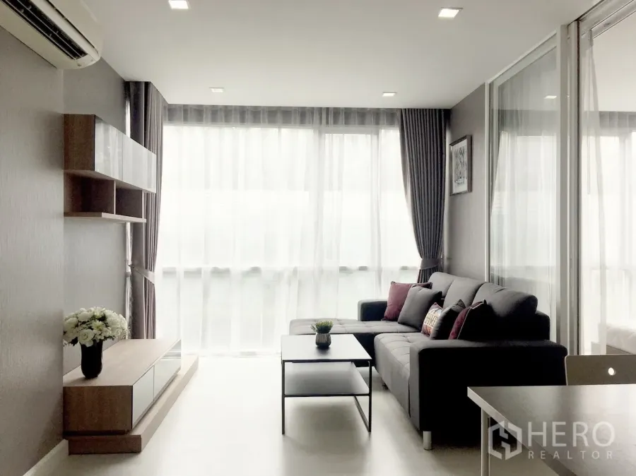 Condominium for sale in Phra Khanong, Bangkok - Bright living room with floor-to-ceiling windows, L-shaped sofa, built-in shelving and a coffee table.