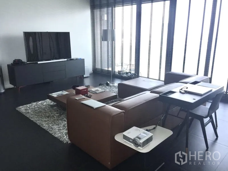 Condominium for rent or sale in Pathum Wan, Bangkok - Modern living room with brown sectional sofa, large TV and floor-to-ceiling windows
