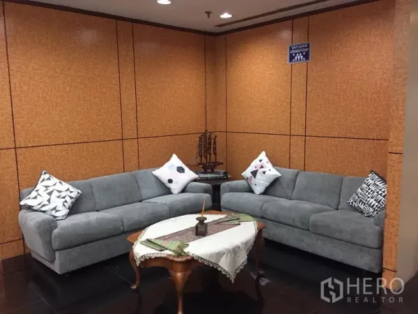 Office Space for rent in Ratchathewi, Bangkok - Corner lounge area featuring two grey sofas, a wooden coffee table with lace runner, and warm wood panel walls under recessed lighting.