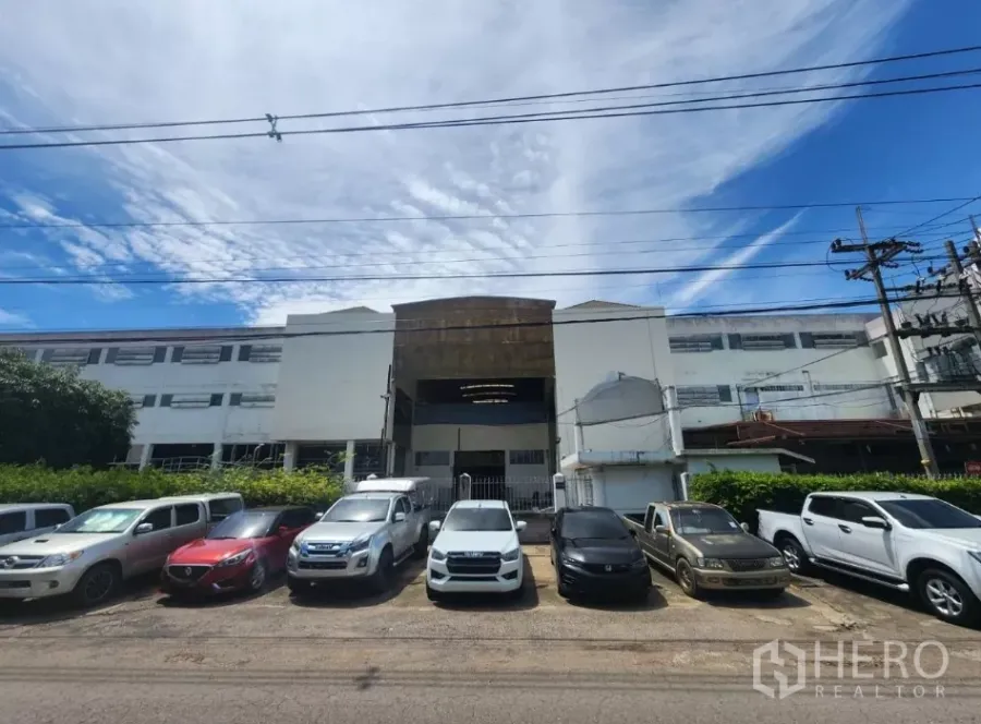 Factory for rent in Mueang Samut Sakhon, Samut Sakhon - Street view of a large multi‑story factory facade with gated entrance and a row of parked cars under a blue sky.