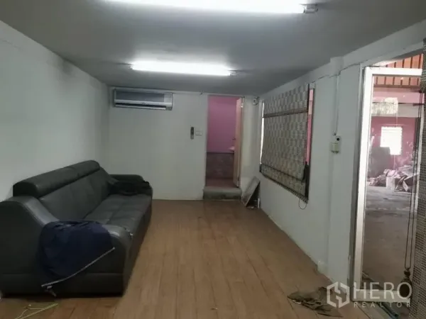Warehouse for rent in Lam Luk Ka, Pathum Thani - Air-conditioned office room with a black sofa, wood-look flooring, fluorescent lights, and window blinds.