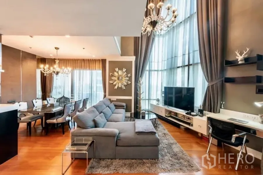 Condominium for rent in Khlong Toei, Bangkok - Expansive living and dining room with tall drapes, chandeliers, and a sectional sofa.
