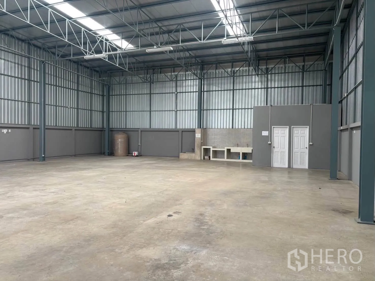 Warehouse for rent in Mueang Pathum Thani, Pathum Thani - Large open warehouse floor with metal cladding, skylight roof panels, sink counter, water tank, and two restroom doors.