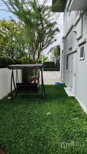 Detached House for rent in Prawet, Bangkok - Side yard with lush lawn, a tree and a garden swing next to the house and white fence.
