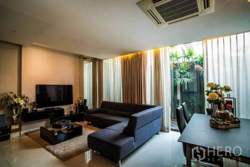 Townhouse for rent or sale in Watthana, Bangkok - Living area with TV wall, glass doors to greenery and an adjoining dining table