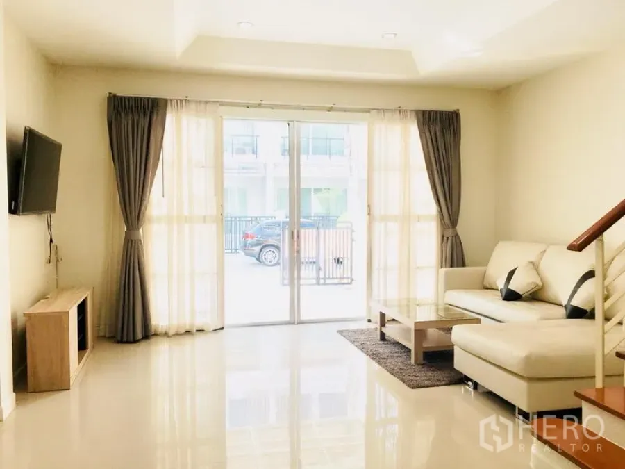 Townhouse for rent in Prawet, Bangkok - Bright living room with L-shaped sofa, wall-mounted TV, and sliding glass doors opening to the front patio and car area.