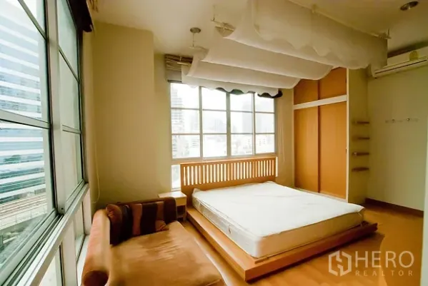 Condominium for rent in Khlong Toei, Bangkok - Sunlit bedroom with wide windows, platform bed, wardrobe and city skyline outlook.