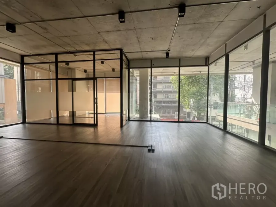 Retail Space for rent - Sunlit open-plan retail space with wood-look flooring, exposed concrete ceiling, and a glass-partitioned room by floor-to-ceiling windows overlooking the street.