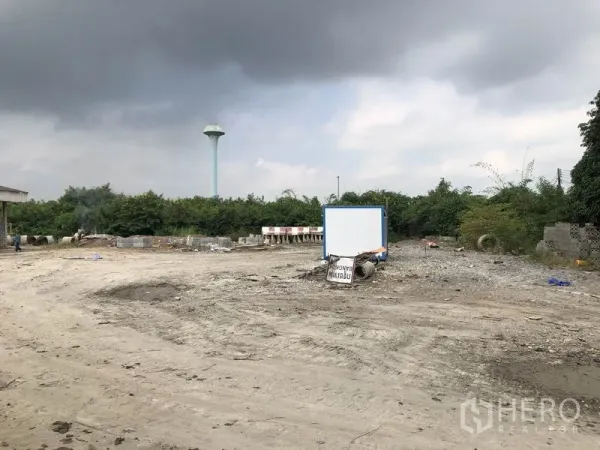 Factory for rent in Phra Nakhon Si Ayutthaya - Open industrial lot with signboard, gravel surface, and a water tower beyond greenery.