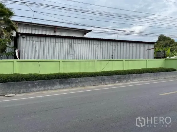 Warehouse for sale in Lam Luk Ka, Pathum Thani - Long corrugated-metal warehouse behind a light green wall along a quiet two-lane road.