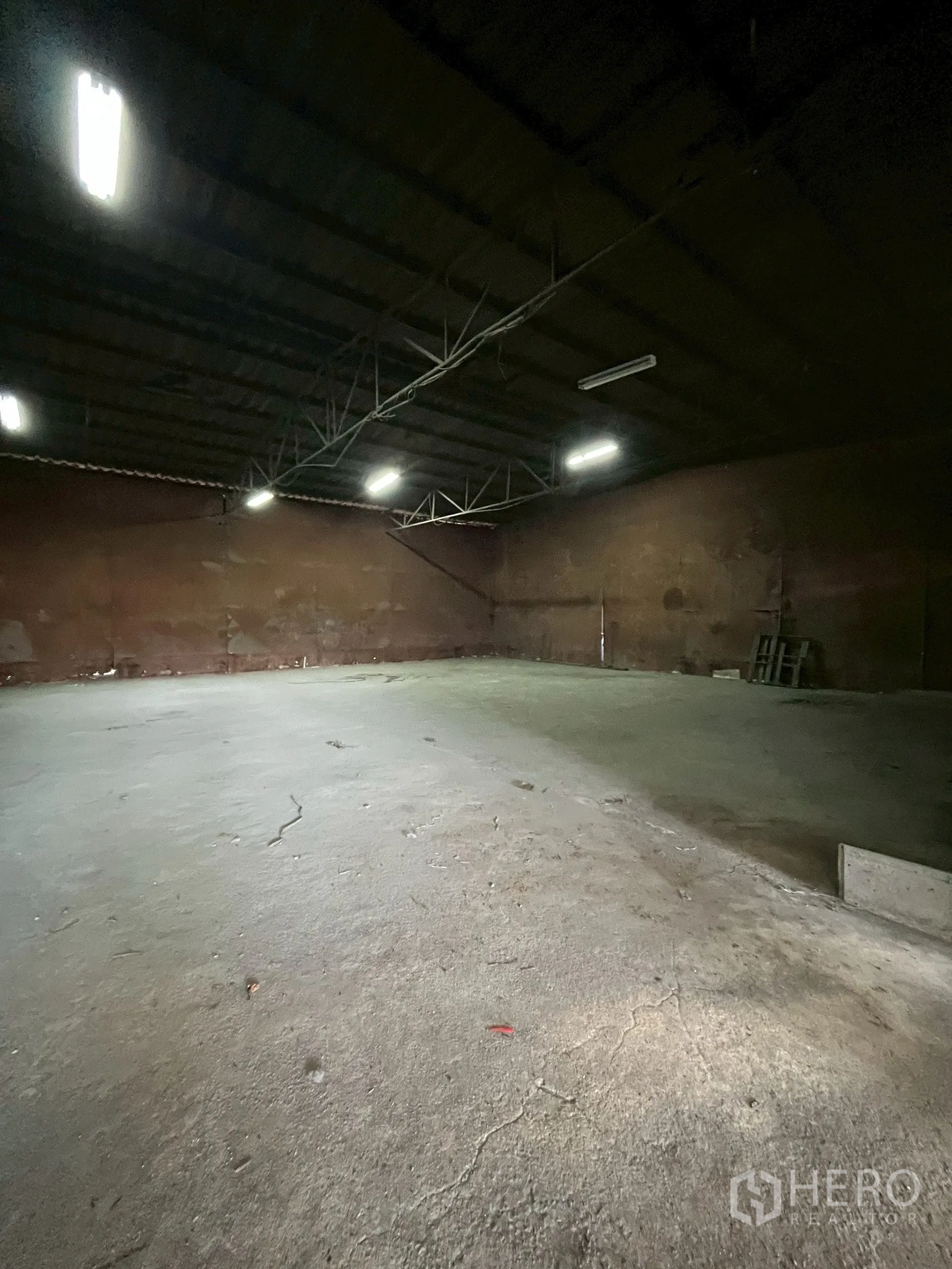 Factory for rent in Phra Pradaeng, Samut Prakan - Interior of the dark storage hall with fluorescent lights and concrete flooring.