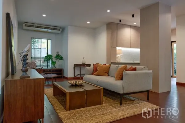 Detached House for rent in Watthana, Bangkok - Warm living room with sofa, rug and built-in kitchen cabinetry under recessed lighting and wood floors.