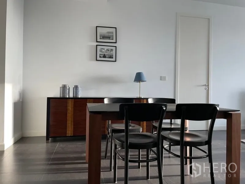 Condominium for rent or sale in Pathum Wan, Bangkok - Dining area with wooden table and four chairs next to a console cabinet and framed artworks.