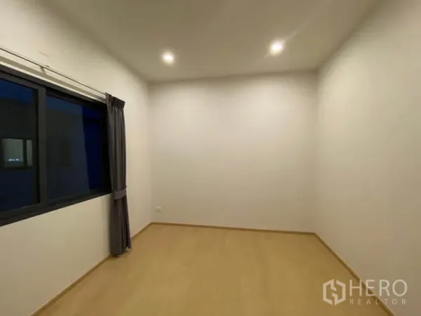 Townhouse for rent in Prawet, Bangkok - Bright empty bedroom with wood-look floor, recessed lights and a window with dark curtains.