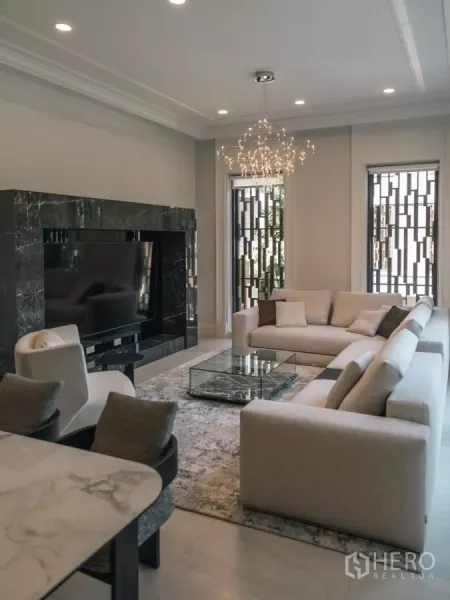 Detached House for rent or sale in Bang Kapi, Bangkok - Elegant living room with cream sofas, marble TV wall, chandelier and tall windows.