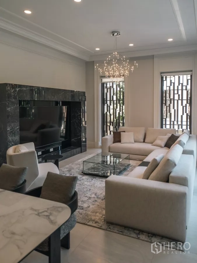 Detached House for rent or sale in Bang Kapi, Bangkok - Elegant living room with cream sofas, marble TV wall, chandelier and tall windows.