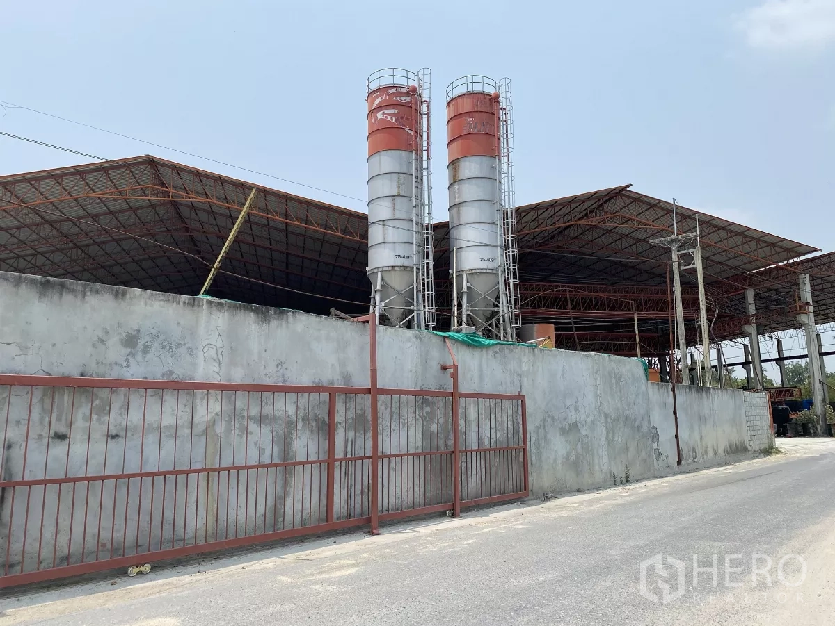 Factory for sale in Chon Buri - Street-side view of the factory wall and gate with two tall cement silos behind.