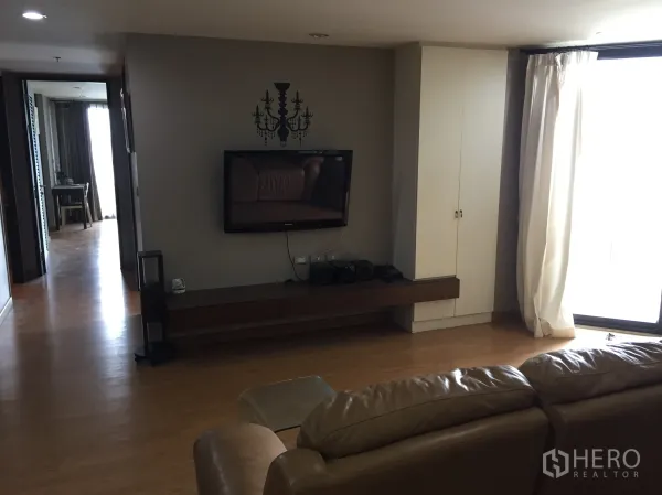 Condominium for rent in Watthana, Bangkok - Living area with wall-mounted TV, long media console, floor-to-ceiling window with curtains, and warm wood flooring.