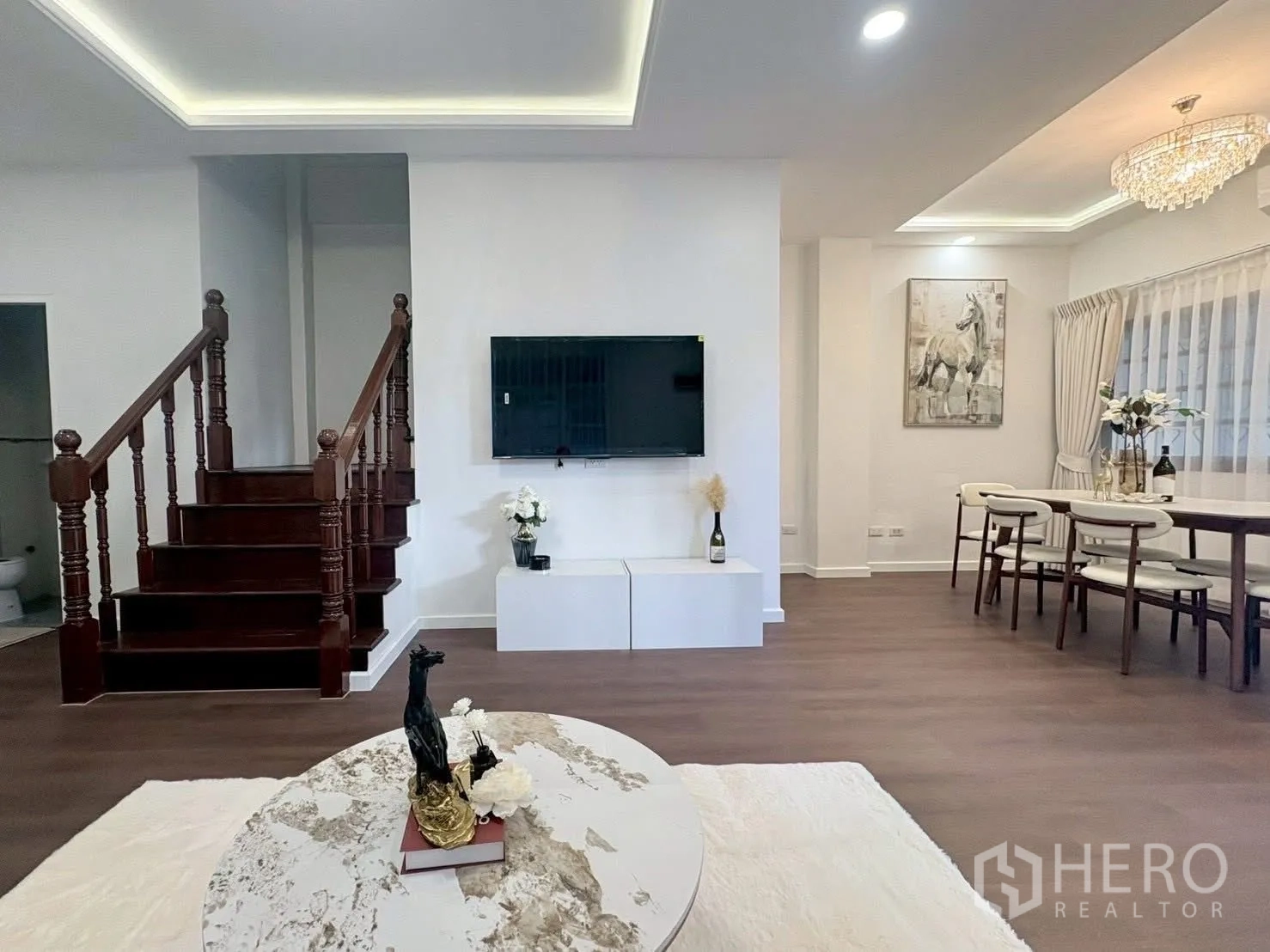 Townhouse for rent in Huai Khwang, Bangkok - Open-plan living and dining area with TV wall, chandelier, and wooden staircase.