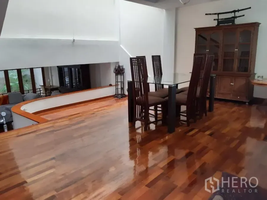 Townhouse for rent in Watthana, Bangkok - Split-level dining area with a glass-top table, high-back wooden chairs, display cabinet, high ceiling and polished hardwood floors.