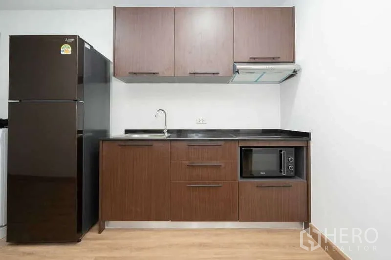 Apartment for rent in Khlong Toei, Bangkok - Modern kitchenette with brown cabinets, black countertop, fridge and microwave.