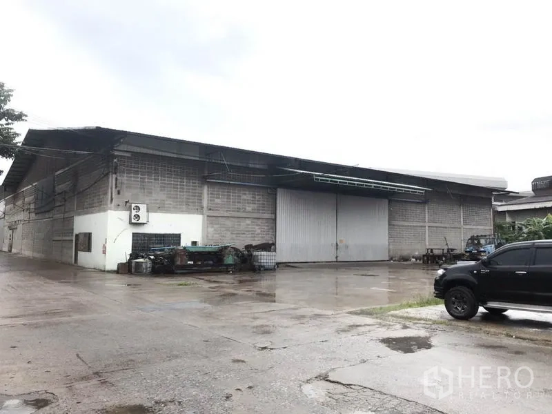 Warehouse for rent in Mueang Pathum Thani, Pathum Thani - Industrial warehouse exterior with large sliding metal doors and a wide concrete yard on a rainy day.