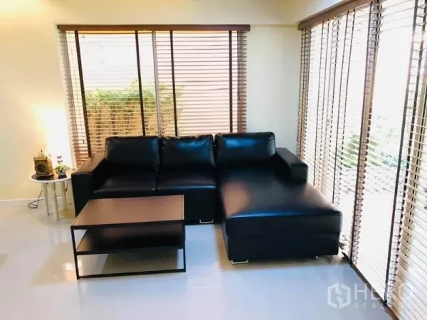 Detached House for rent in Watthana, Bangkok - Corner living space with wraparound windows, blinds, a black sectional sofa, and a coffee table.