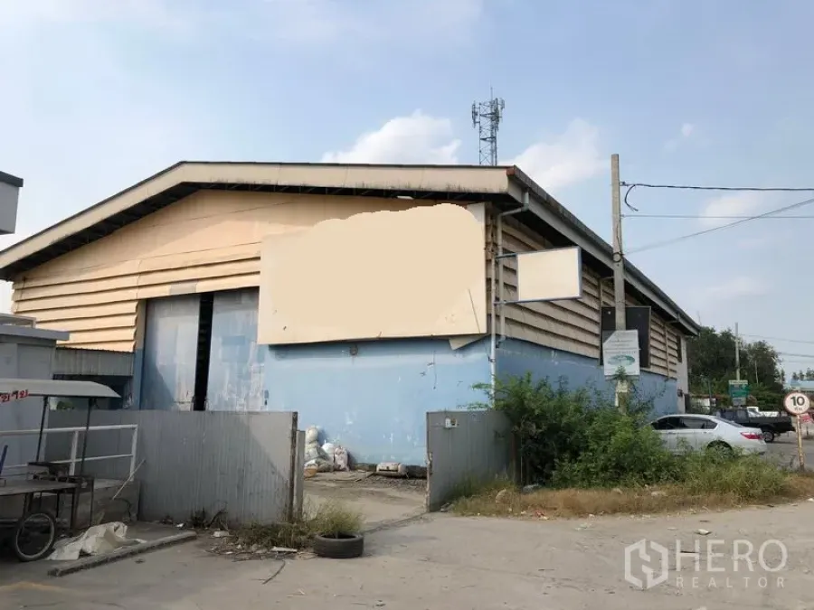 货栈 出租在Nonthaburi - 仓库外观，山墙屋顶与蓝色外立面，带铁门与院落，临路电线杆。