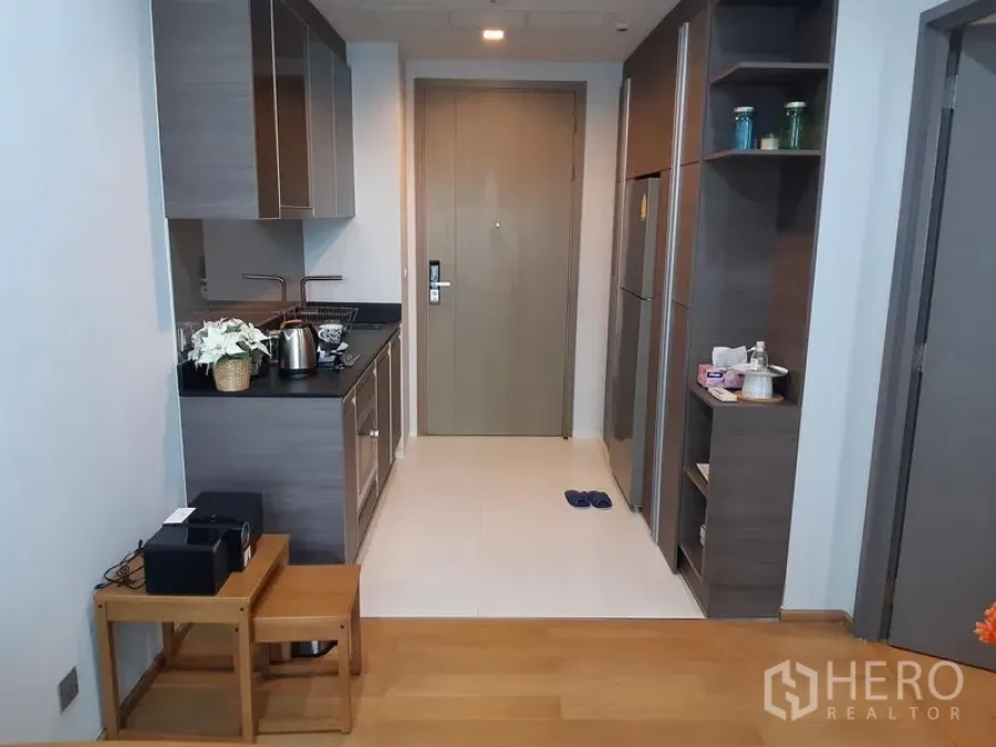 Condominium for rent in Khlong Toei, Bangkok - Compact galley kitchen with stone countertop, upper cabinets, sink, cooktop and tall fridge beside the entry door.