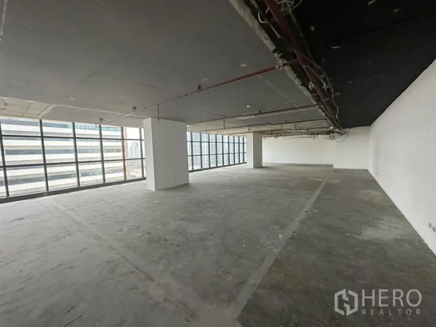 Office Space for rent - Expansive bare concrete office space with structural columns and full-height windows along two sides.