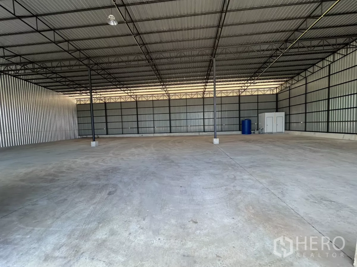 Warehouse for rent in Ban Pho, Chachoengsao - Wide warehouse interior with high ceiling, steel columns, concrete floor, and a small restroom unit and water tank at the rear.