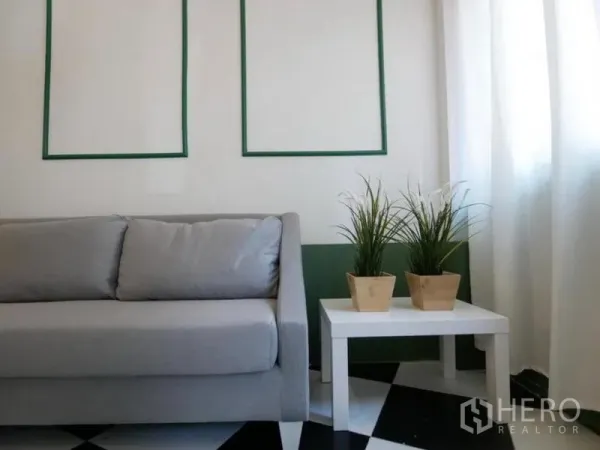 Townhouse for rent in Khlong Toei, Bangkok - Close-up of a gray sofa beside a white side table with potted plants, sheer curtains and a monochrome tiled floor.