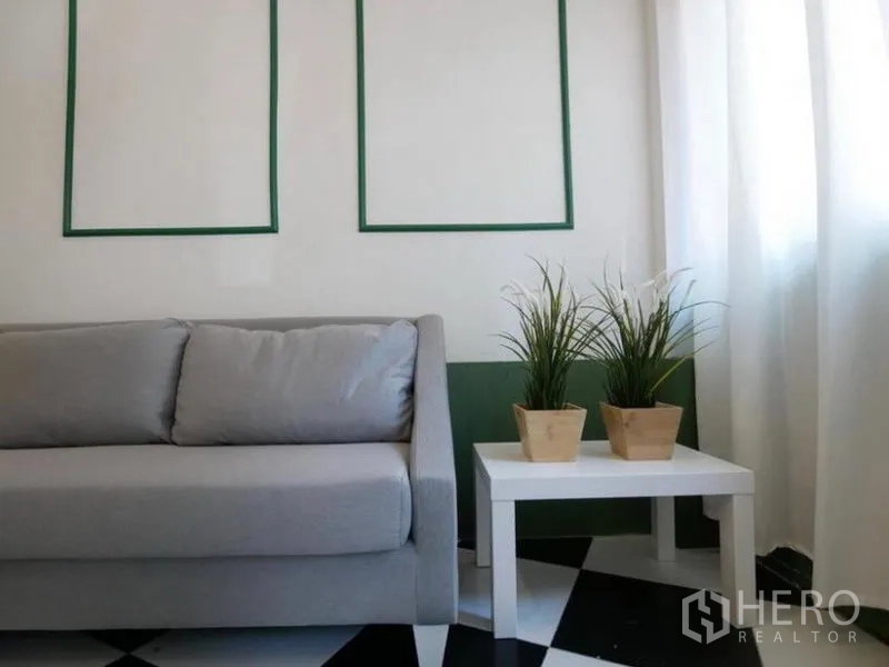 Townhouse for rent in Khlong Toei, Bangkok - Close-up of a gray sofa beside a white side table with potted plants, sheer curtains and a monochrome tiled floor.