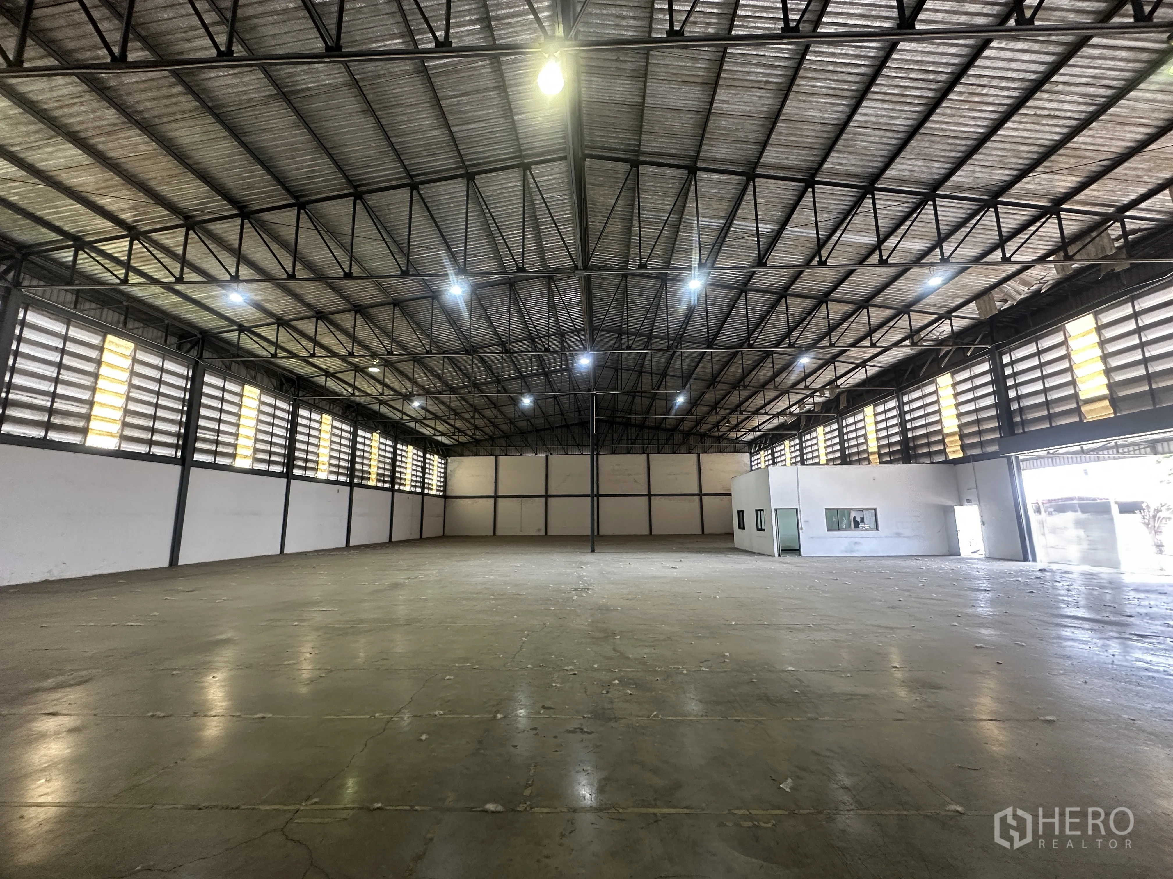 Warehouse for rent in Khlong Luang, Pathum Thani - Open concrete floor area with bright lighting, louvered windows, and an office section on the side.