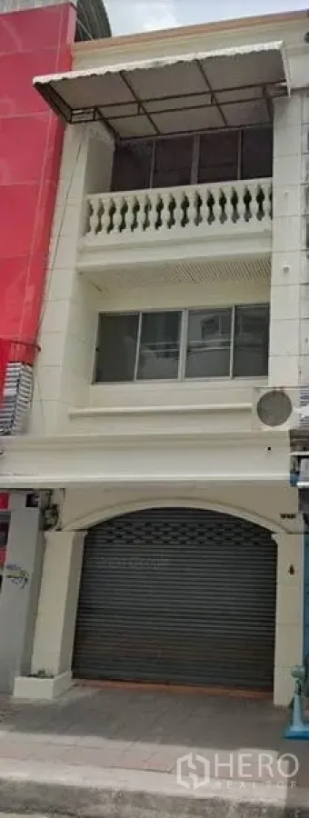 Shophouse for rent in Watthana, Bangkok - Three-story white shophouse facade with closed roll-up shutter, balcony, and awning on a city street.