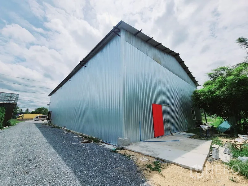 Warehouse for rent in Sam Phran, Nakhon Pathom - Side exterior of corrugated metal warehouse with a bright red door and service walkway.