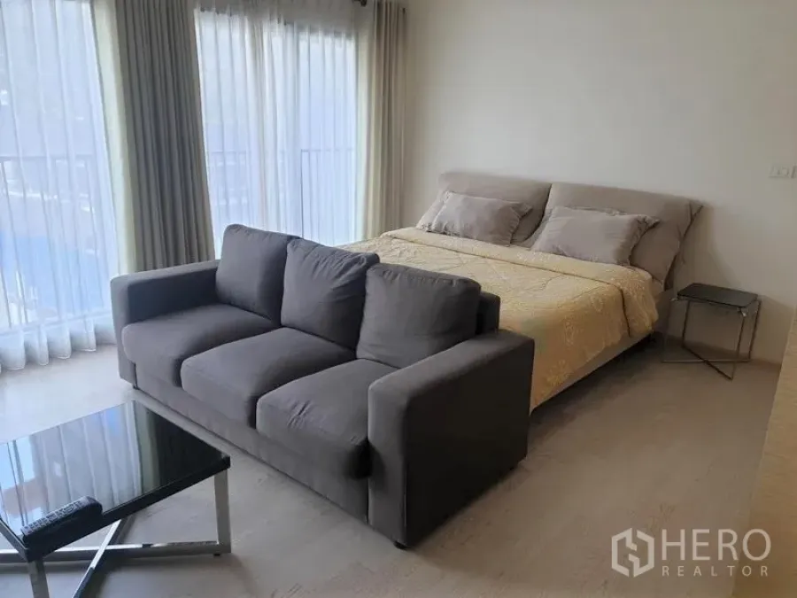 Condominium for rent in Khlong Toei, Bangkok - Bright sleeping area with large bed, gray sofa and corner floor-to-ceiling windows with sheer curtains.