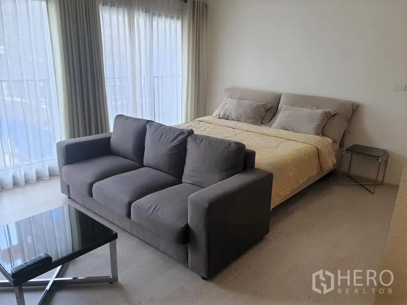 Condominium for rent in Khlong Toei, Bangkok - Bright sleeping area with large bed, gray sofa and corner floor-to-ceiling windows with sheer curtains.