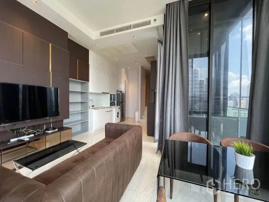 公寓 出租在Bang Rak, Bangkok - 开放式客餐厨布局，棕色沙发与电视墙，转角玻璃窗展示城市景观。