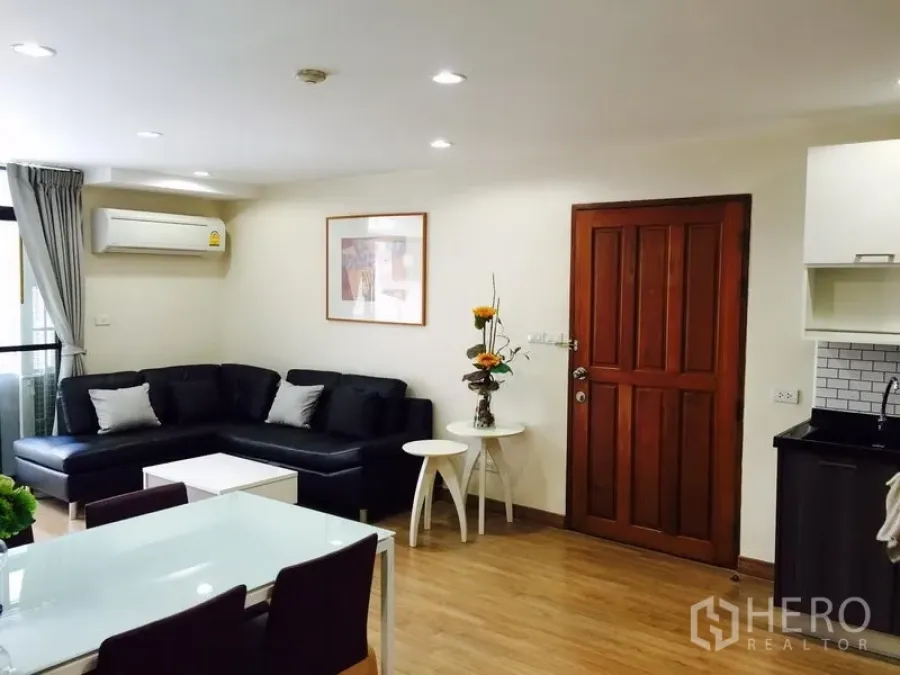 Condominium for rent in Watthana, Bangkok - Bright living and dining area with black L-shaped sofa, wood floors, and air conditioner.