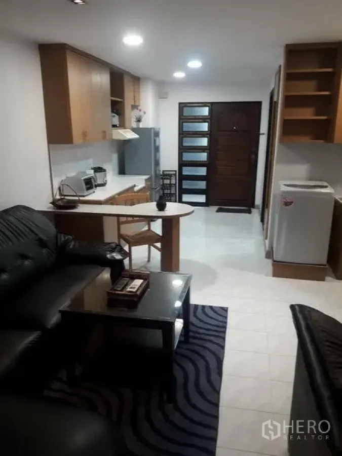 Condominium for rent in Watthana, Bangkok - Open-plan living room with black leather sofa, coffee table, kitchen and in-unit washer.