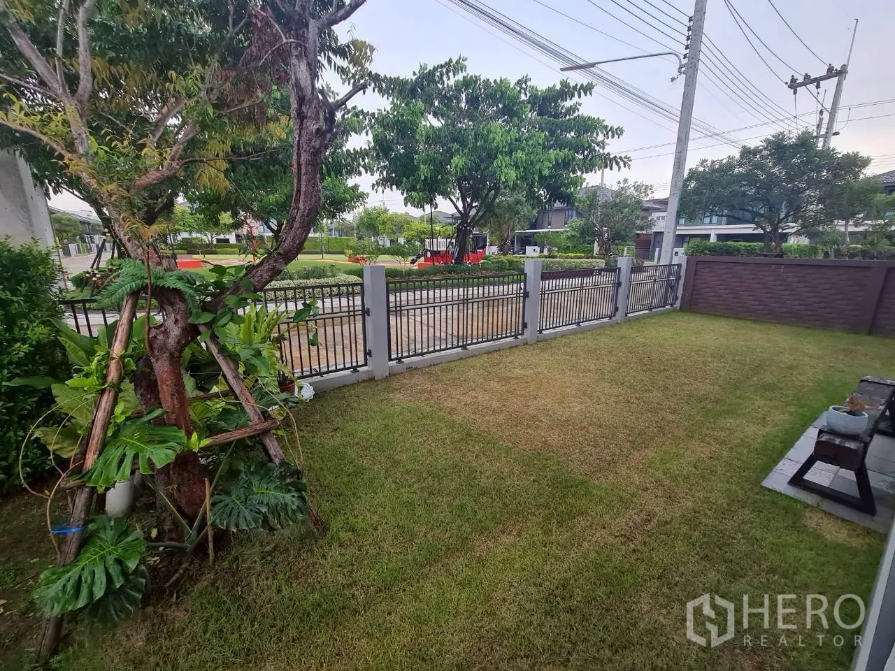 Detached House for rent or sale in Lam Luk Ka, Pathum Thani - Front garden lawn with trees and a metal fence facing the community road.