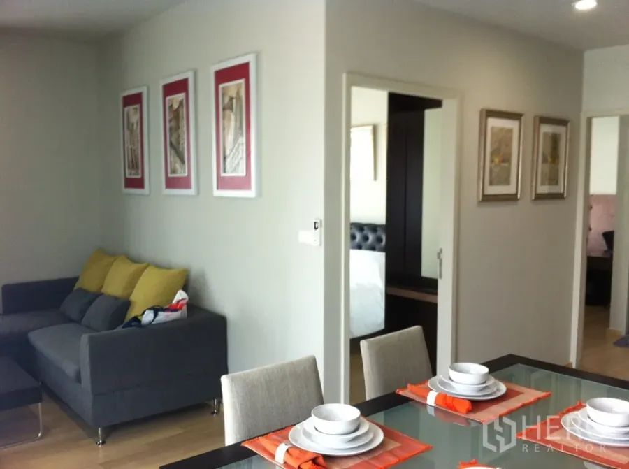 Condominium for rent in Bangkok - Dining area with glass-top table and place settings beside a gray sofa with yellow cushions, leading toward a bedroom with a mirrored wardrobe.